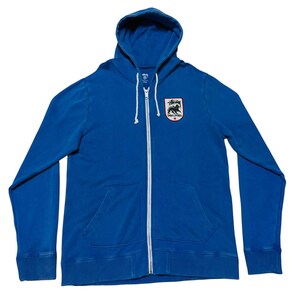 May include: A blue zip-up hoodie with a hood and a kangaroo pocket. The hoodie features a white zipper and a patch on the chest with a black and white graphic and the text "STUSSY". The hoodie is made of a soft material.