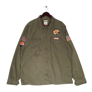 May include: A green long-sleeved button-down shirt with multiple patches, including a tiger, a mountain, and an American flag. The shirt is made of a cotton blend and has a relaxed fit.