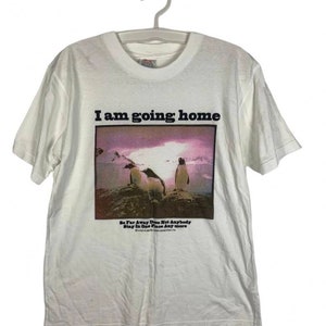 May include: White t-shirt with a graphic of three penguins on a rocky surface under a purple sky. The text "I am going home" is above the graphic, with additional text below.