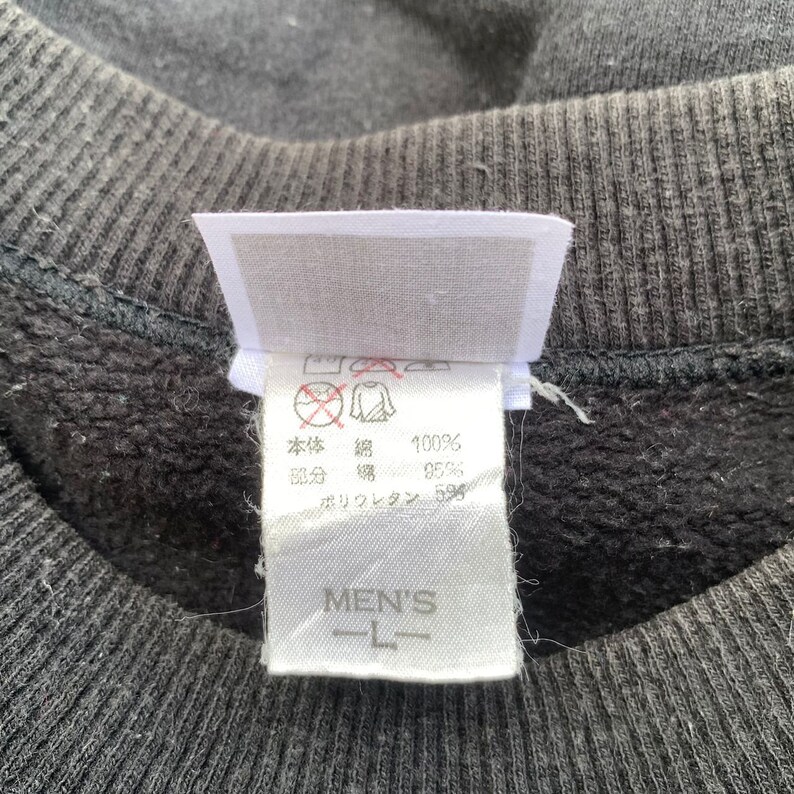 K&ouml;nnte beinhalten: Ein wei&szlig;es Etikett mit schwarzem Text, das die Stoffzusammensetzung eines schwarzen Sweatshirts beschreibt. Das Etikett lautet "K&ouml;rper: 100 % Baumwolle, Rippen: 95 % Baumwolle, 5 % Elasthan". Das Etikett enth&auml;lt auch ein Pflegeetikett mit Symbolen f&uuml;r Waschen, Trocknen und B&uuml;geln. Das Etikett ist mit "Men's L" gekennzeichnet.