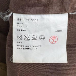 May include: A clothing label with Japanese text and symbols, featuring the product code "TL-C005" and size "S". The label is white with black text and a red stamp. The background is a brown fabric, likely part of the garment.