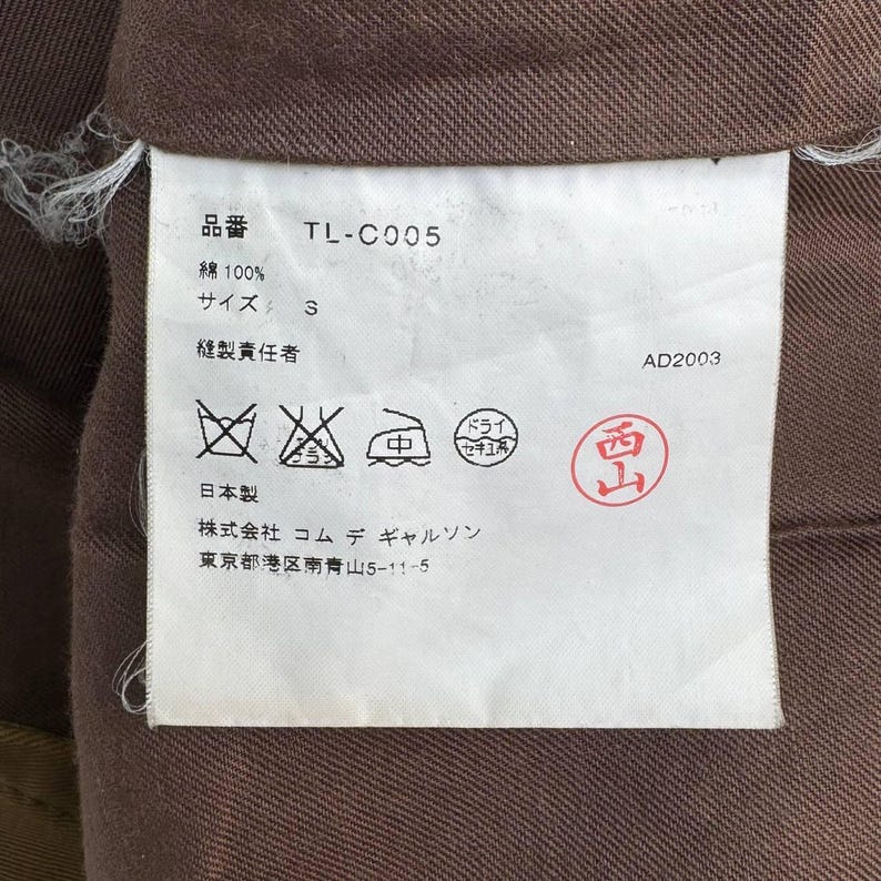 May include: A clothing label with Japanese text and symbols, featuring the product code "TL-C005" and size "S". The label is white with black text and a red stamp. The background is a brown fabric, likely part of the garment.