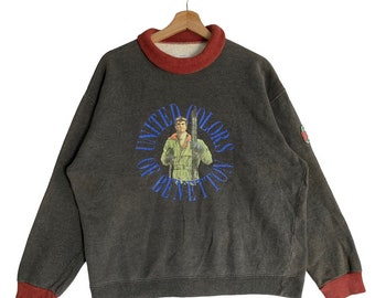 Puede incluir: Una sudadera gris oscuro con cuello y puños de canalé rojos. La parte delantera de la sudadera presenta un logotipo circular con el texto "United Colors of Benetton" en azul y un gráfico de un hombre esquiando.