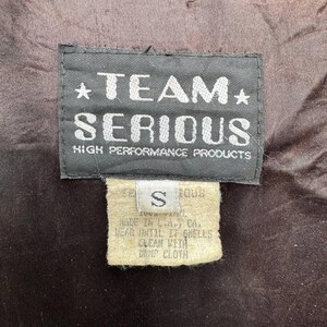 Op de afbeelding: Een zwart-wit label met de tekst "*TEAM* SERIOUS HIGH PERFORMANCE PRODUCTS" en een kleiner wit label met de tekst "S" en wasinstructies.