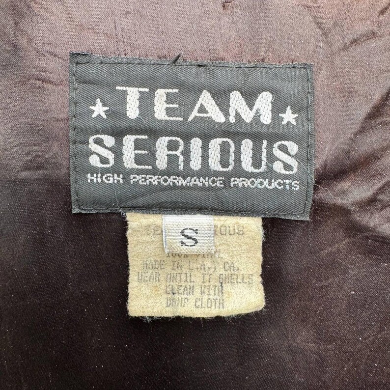 Op de afbeelding: Een zwart-wit label met de tekst "*TEAM* SERIOUS HIGH PERFORMANCE PRODUCTS" en een kleiner wit label met de tekst "S" en wasinstructies.