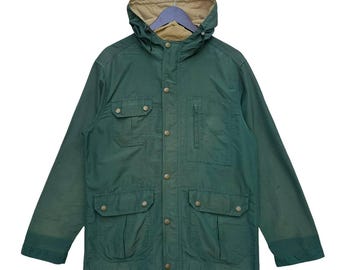 May include: A green hooded jacket with a tan-colored hood lining. The jacket has multiple pockets, button closures, and a drawstring hood. The jacket is hanging on a hanger.