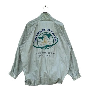 May include: A light green oversized jacket with a high collar and a zipper closure. The back of the jacket features a circular logo with the words "WORLD STAGE" and "OVERSIZED METAL". The logo includes the letters "Js" and a globe.