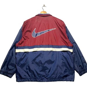 Op de afbeelding: Een rood en blauw windjack met een witte streep en een groot Nike swoosh-logo op de rug.