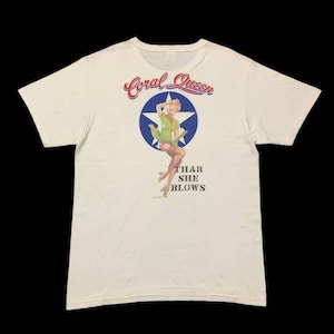 May include: White t-shirt with a graphic of a pin-up girl in a green dress and red heels standing on a blue star with the text "Coral Queen" in red and "Thar She Blows" in black.