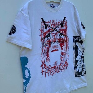 May include: White t-shirt with a graphic print of a red and black skull with crossed skateboards and the text "Don't mess with Texas". The shirt also has a graphic print of a black and white skull on the right sleeve and a blue and white graphic print on the left sleeve.