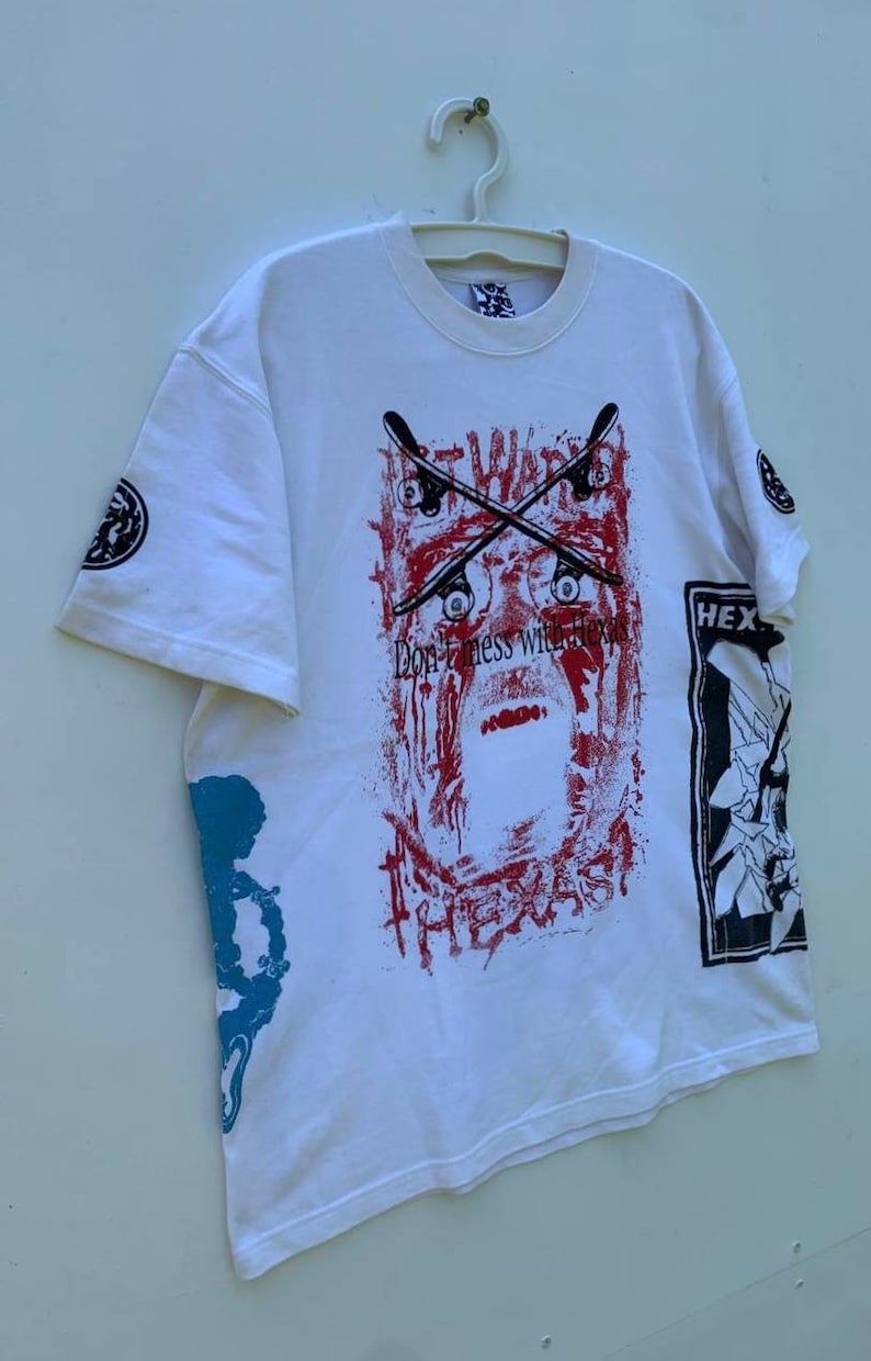 May include: White t-shirt with a graphic print of a red and black skull with crossed skateboards and the text "Don't mess with Texas". The shirt also has a graphic print of a black and white skull on the right sleeve and a blue and white graphic print on the left sleeve.