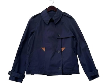 Puede incluir: Chaqueta azul marino con un diseño clásico. La chaqueta presenta un cuello, cierres de botones y dos bolsillos delanteros con detalles de cuero marrón. Las mangas tienen puños abotonados. La chaqueta está colgada de una percha de madera.