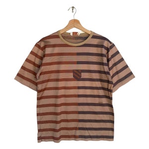 May include: A brown and tan striped short-sleeved t-shirt with a brown and tan striped pattern. The shirt has a small logo on the left side and the word "GUCCI" on the right side.