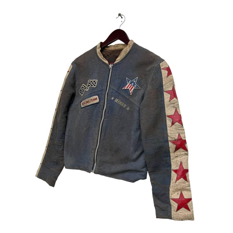Op de afbeelding: Een blauwe denim jas met rode en witte ster-patches op de mouwen en een raceteam-patch op de voorkant. De jas heeft een ritssluiting en een opstaande kraag.