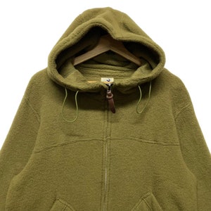 K&ouml;nnte beinhalten: Ein gr&uuml;ner Fleece-Kapuzenpullover mit braunem Rei&szlig;verschluss und einer Kapuze mit Kordelzug.