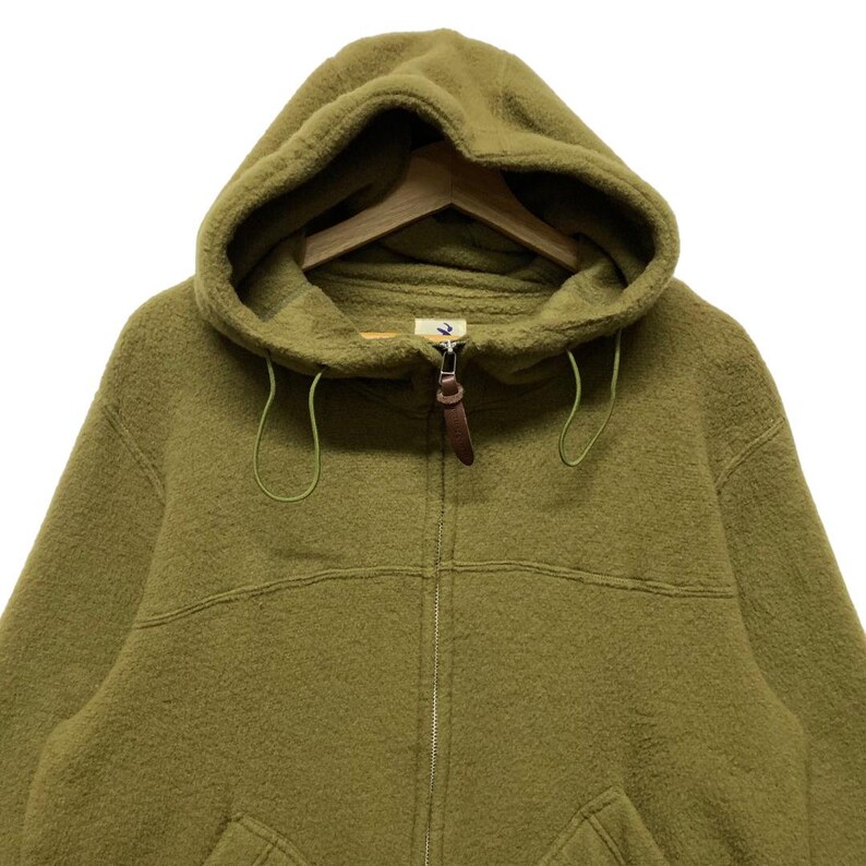 K&ouml;nnte beinhalten: Ein gr&uuml;ner Fleece-Kapuzenpullover mit braunem Rei&szlig;verschluss und einer Kapuze mit Kordelzug.