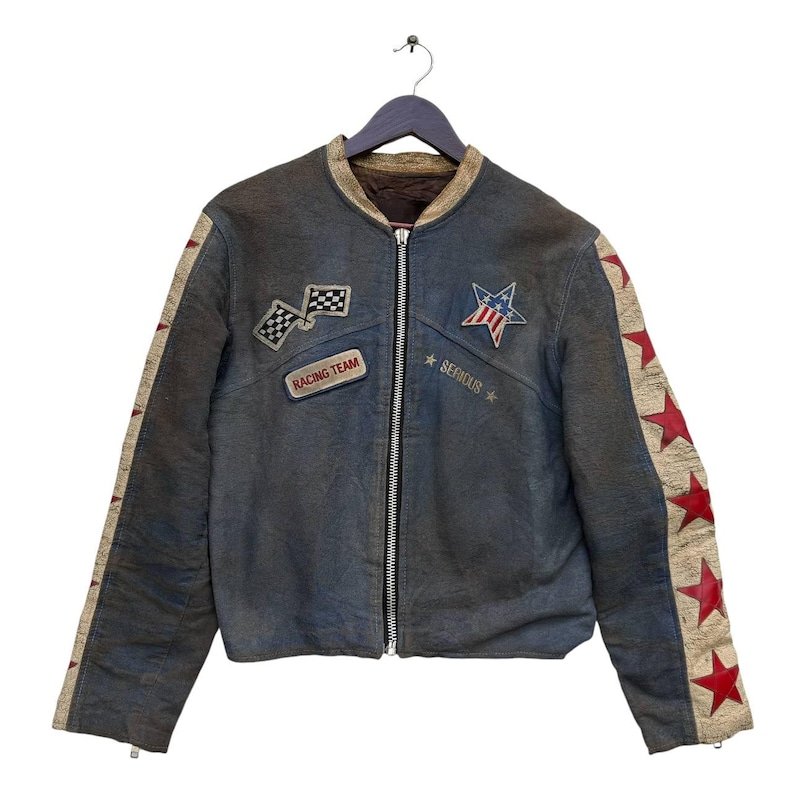 Op de afbeelding: Een blauw leren jack met rode en witte ster-patches op de mouwen en een raceteam-patch op de voorkant. Het jack heeft een ritssluiting en een opstaande kraag.