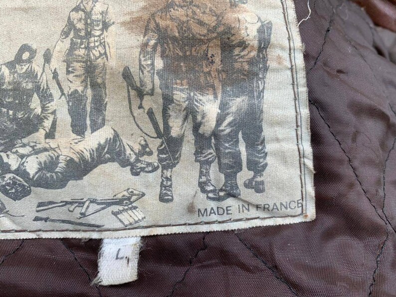 May include: A close-up of a brown leather jacket with a white label sewn into the inside. The label features a black and white illustration of soldiers and the text "Made in France".