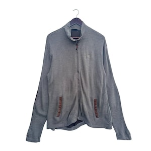 Puede incluir: Chaqueta gris con cremallera y cuello alto, colgada de una percha de madera. La chaqueta tiene dos bolsillos delanteros con detalles de cuero marrón. Un pequeño logotipo es visible en el pecho. Las mangas son largas y los puños oscuros.