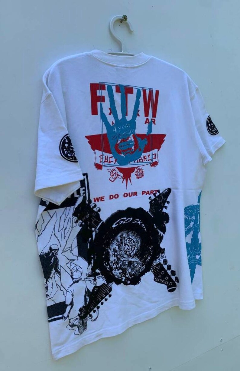 May include: White t-shirt with a graphic print of a hand with the text "FTW" and "4 year anniversary" and "FUCK THE WORLD". The shirt also has a graphic print of guitars and the text "WE DO OUR PART" and "GO SKATE".