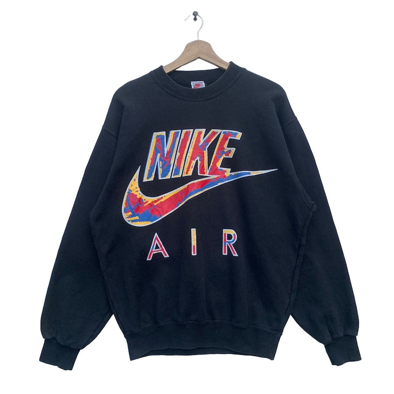 K&ouml;nnte beinhalten: Schwarzer Nike Air Sweatshirt mit einem bunten, abstrakten Nike Swoosh Logo und dem Wort "NIKE" in einer fetten, bunten Schrift.