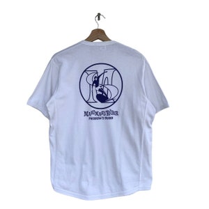 May include: White short-sleeved t-shirt with a blue graphic of a person surfing on a wave inside a circle with the text "MAHI MAHI RIDER PHERROW'S BRAND" underneath.