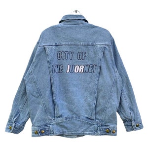 May include: A light blue denim jacket with a white background and the text "CITY OF THE JOURNEY" embroidered on the back.