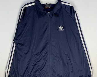 May include: A navy blue zip-up jacket with white stripes on the sleeves and a white Adidas logo on the chest. The jacket has a ribbed collar and cuffs.