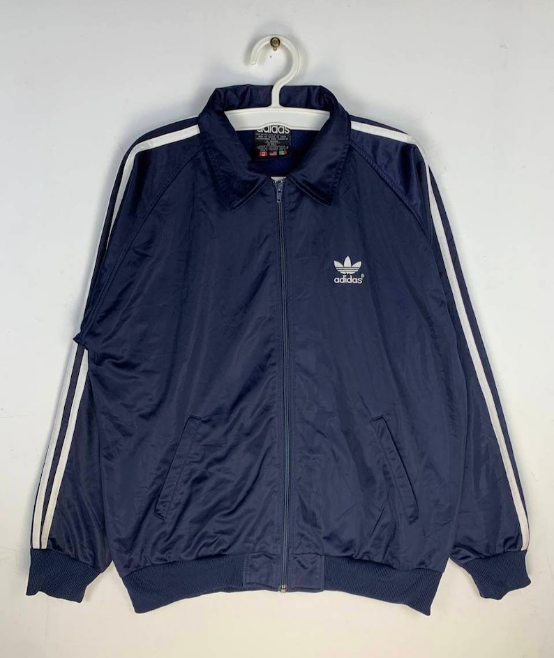 May include: A navy blue zip-up jacket with white stripes on the sleeves and a white Adidas logo on the chest. The jacket has a ribbed collar and cuffs.