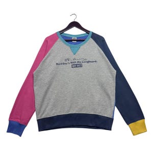 May include: A gray sweatshirt with a colorful color-blocked design. The sweatshirt has a crew neck and long sleeves. The sleeves are pink, blue, and yellow. The text "Surf Day's With His Longboard. Surf Day's" is printed on the front of the sweatshirt.