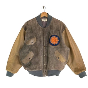 Könnte beinhalten: Eine braune Wildleder-Bomberjacke mit Lederärmeln. Die Jacke hat ein "BIGI"-Etikett auf der Vorderseite und einen Patch mit einer Sonne und dem Buchstaben "B" auf der linken Seite.