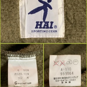 K&ouml;nnte beinhalten: Nahaufnahme eines gr&uuml;nen Stofflabels mit wei&szlig;em Text, der "HAI SPORTING GEAR" lautet. Das Etikett zeigt au&szlig;erdem eine blaue Silhouette einer Person, die einen Karate-Tritt ausf&uuml;hrt. Das Etikett ist an einem gr&uuml;nen Kleidungsst&uuml;ck befestigt.