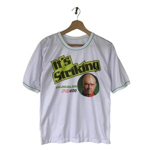 Puede incluir: Camiseta blanca con un gráfico verde y negro que dice "It's Striking" y una foto de un hombre. El gráfico también incluye el texto "Fujicolor F-II 400".