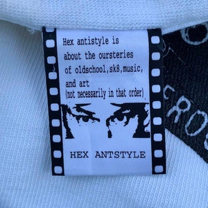 May include: White clothing tag with black text that reads "Hex antistyle is about the oursteries of oldschool, sk8, music, and art (not necessarily in that order)" and "HEX ANTSTYLE". The tag features a black and white illustration of two eyes with a single teardrop.