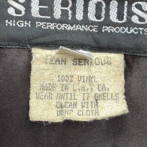 Op de afbeelding: Een wit verzorgingsetiket met zwarte tekst die luidt: "Team Serious 100% Vinyl Made in L.A. CA. Wear until it smells Clean with damp cloth"