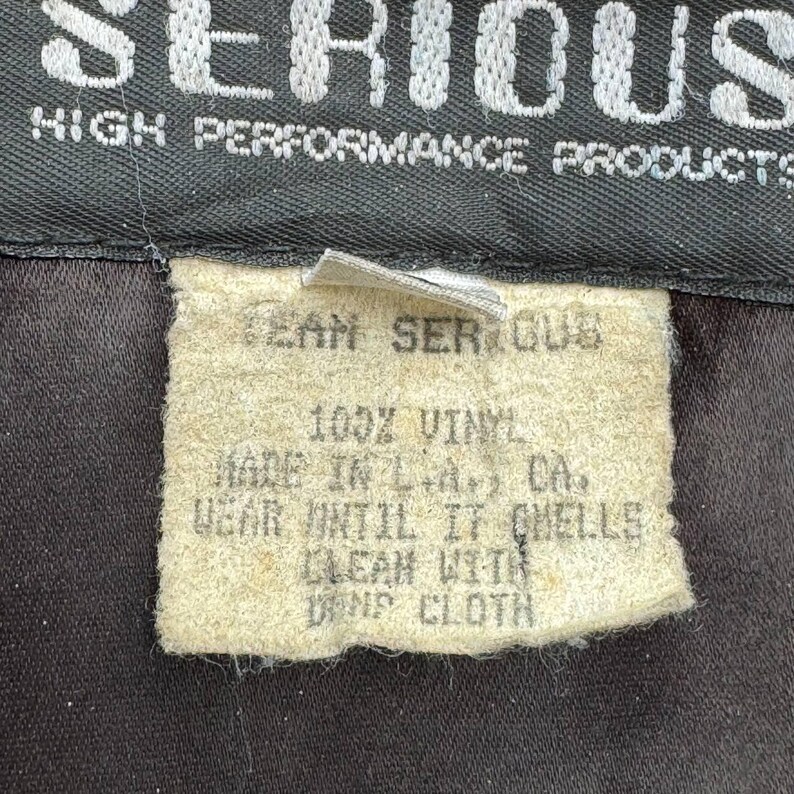Op de afbeelding: Een wit verzorgingsetiket met zwarte tekst die luidt: "Team Serious 100% Vinyl Made in L.A. CA. Wear until it smells Clean with damp cloth"