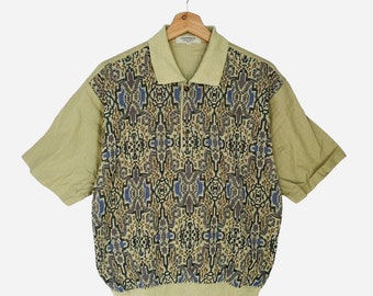 May include: A short-sleeved, light green polo shirt with a geometric pattern in shades of brown, blue, and beige. The shirt has a button-up collar and a zipper closure.