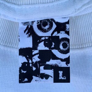 May include: White t-shirt with a black and white graphic print of eyes and the letter "L" on a tag.
