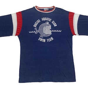 May include: A blue short-sleeved t-shirt with red and white striped sleeves. The shirt has a white graphic of a fish with the text "Racine Aquatic Club Swim Team".
