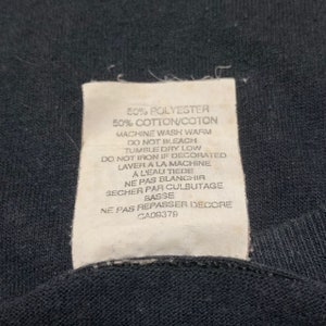 May include: White care label with black text detailing the fabric composition and care instructions for a garment. The label reads: 50% Polyester 50% Cotton/Coton Machine Wash Warm Do Not Bleach Tumble Dry Low Do Not Iron If Decorated Laver a la Machine a l'eau tiede Ne Pas Blanchir Secher par Culbutage Basse Ne Pas Repasser Decore CA08378