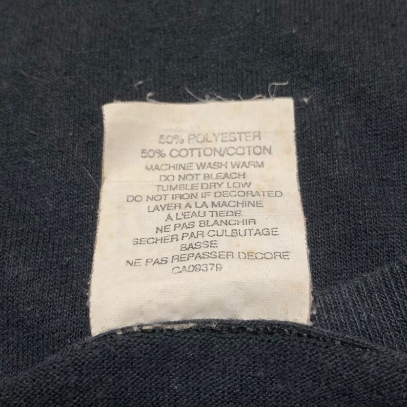 May include: White care label with black text detailing the fabric composition and care instructions for a garment. The label reads: 50% Polyester 50% Cotton/Coton Machine Wash Warm Do Not Bleach Tumble Dry Low Do Not Iron If Decorated Laver a la Machine a l'eau tiede Ne Pas Blanchir Secher par Culbutage Basse Ne Pas Repasser Decore CA08378