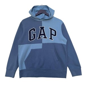 May include: A blue hooded sweatshirt with a large "GAP" logo in a navy blue color. The sweatshirt has a patchwork design with different shades of blue.