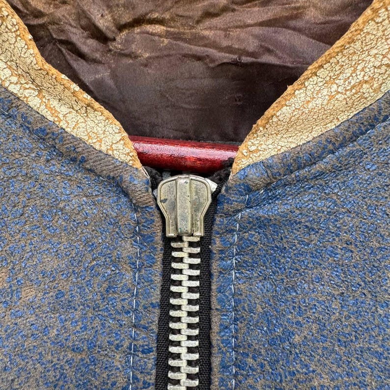Op de afbeelding: Close-up van een blauw leren jack met een zilveren rits. Het jack heeft een bruine leren kraag en is verweerd.