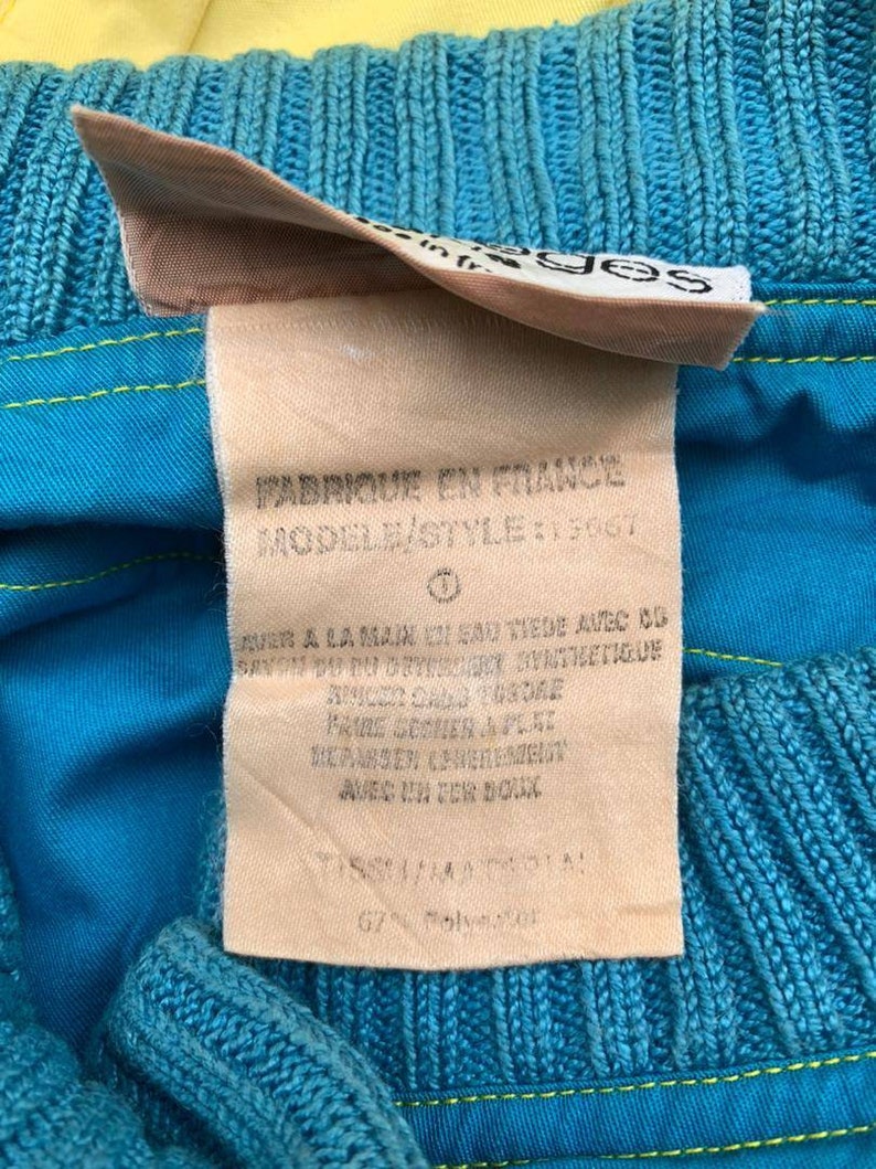 May include: A close-up of a blue and yellow fabric label with the text "FABRIQUE EN FRANCE" and "MODELE/STYLE: 13067" in black. The label also includes care instructions in French.