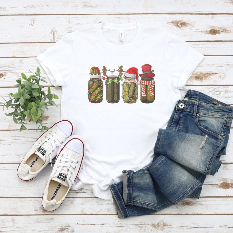 May include: White t-shirt with a graphic of four pickle jars decorated for Christmas. Each jar has a different festive hat and is adorned with lights and greenery.