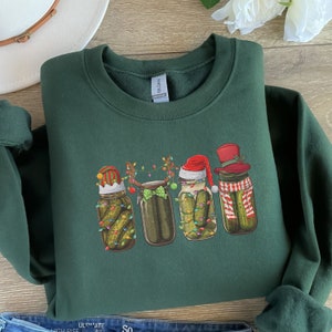 May include: A dark green sweatshirt with a Christmas-themed design featuring four pickle jars decorated with festive hats, lights, and bows.