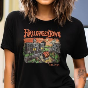 May include: Black t-shirt with a graphic print of a spooky town with a large pumpkin in the center. The text "Halloween Town Est. 1998" is printed below the graphic.
