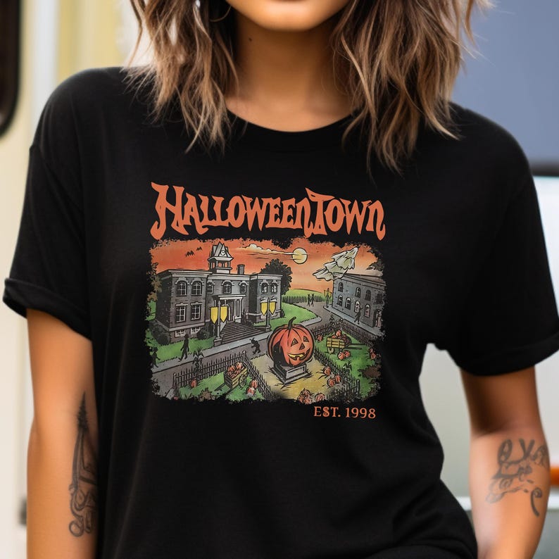 May include: Black t-shirt with a graphic print of a spooky town with a large pumpkin in the center. The text "Halloween Town Est. 1998" is printed below the graphic.