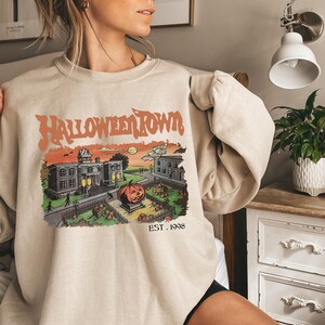 May include: A beige sweatshirt with a graphic print of a spooky town with a pumpkin and the text "HALLOWEEN TOWN EST. 1998".