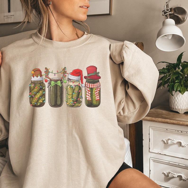 May include: A beige sweatshirt with a graphic of four pickle jars decorated for Christmas. Each jar has a different festive hat and is adorned with lights and other holiday decorations.